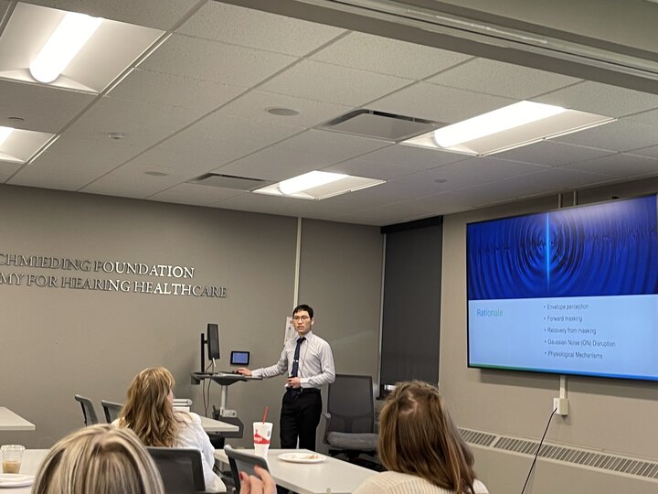 A young man presenting in a modern conference room with a large screen displaying the title of his capstone project.