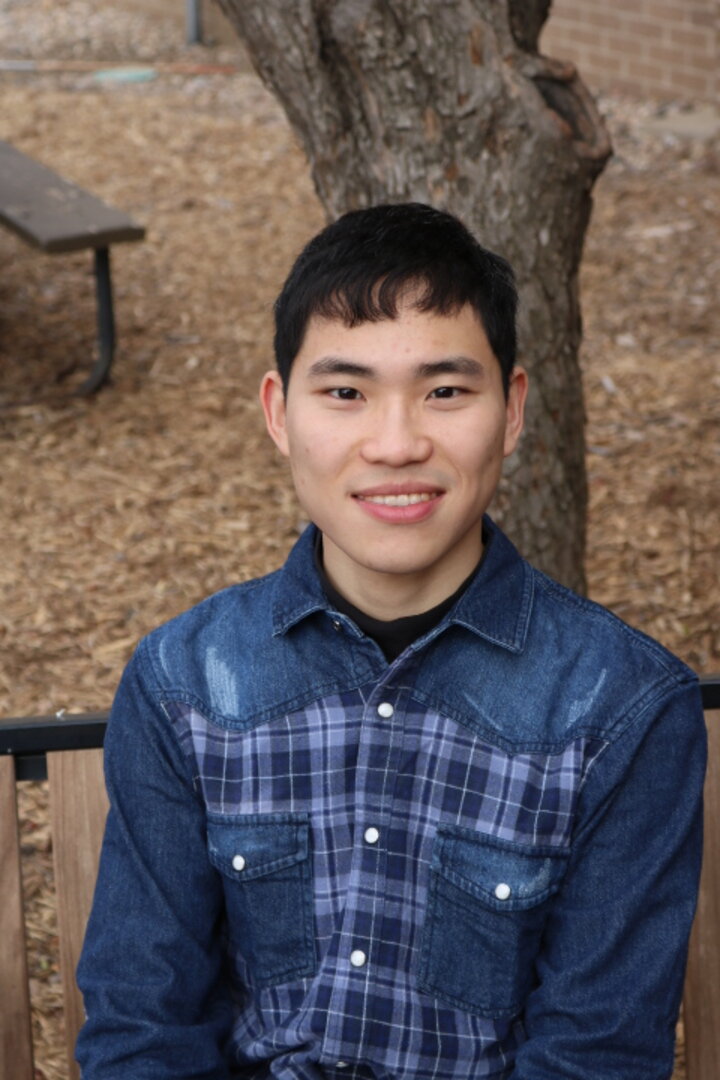 Cheng-Han outside on a bench with dry fallen leaves and a tree in the background. He is wearing a blue plaid shirt with white stripes.