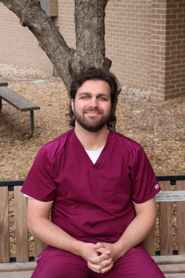 Sam Aguilar  on a bench with dry fallen leaves and a tree in the background. He's wearing rose colored scrubs.